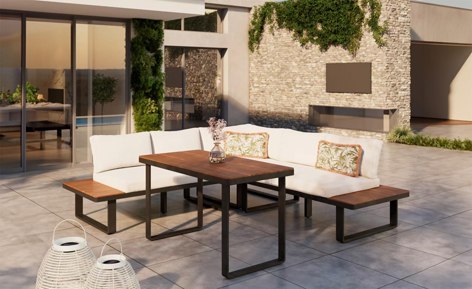 Modern teraszon fehér párnázott sarokkanapé fa asztallal és padokkal; háttérben kőburkolatú fal futónövénnyel, az előtérben fehér lámpások