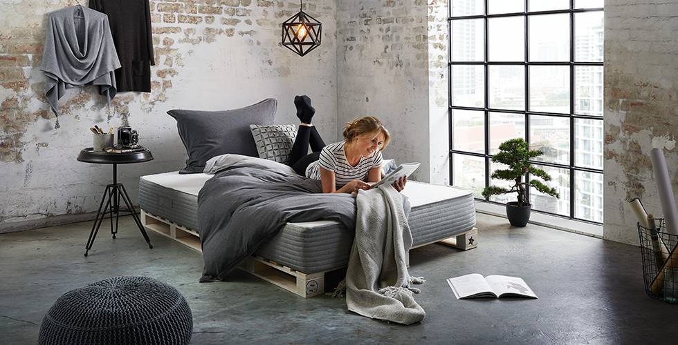 Palettenbett und dunkelgrau Boxspringmatratze von mömax, bunt mit Decken und Kissen dekoriert.