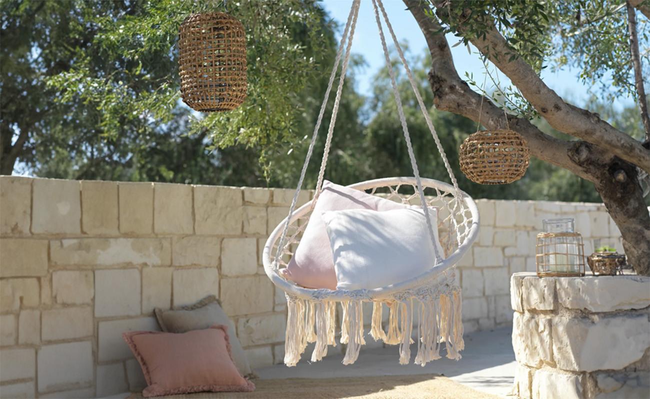 Fehér, rojtos függőszék párnákkal egy árnyas kertben; a fa ágairól fonott lámpások lógnak, háttérben világos kőfal és kültéri asztalka dekorációval.
