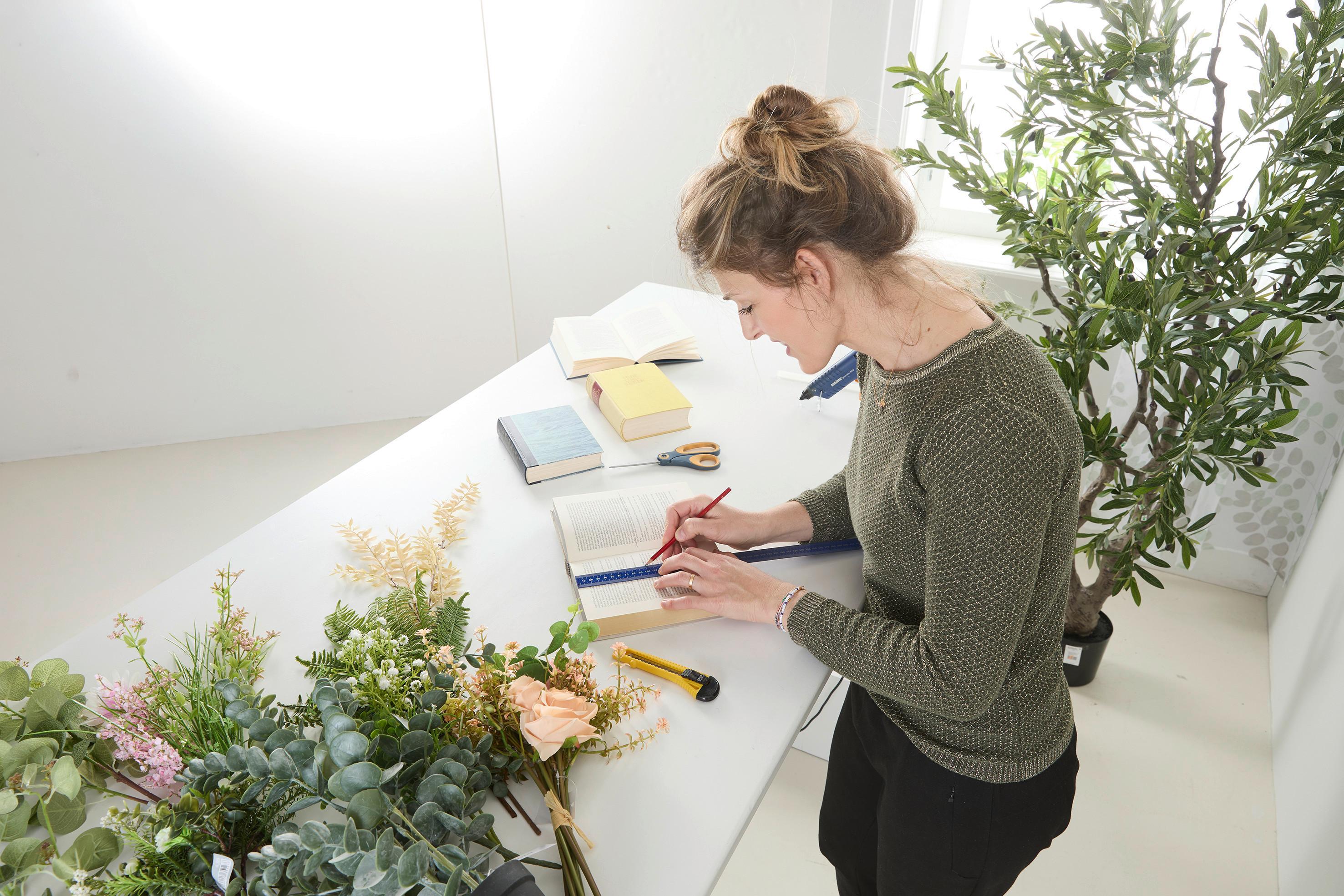 Asztalnál dolgozó nő egy könyv lapjait méri és jelöli vonalzóval és ceruzával; mellette olló, sniccer, ragasztópisztoly és virágok egy DIY könyvváza elkészítéséhez.