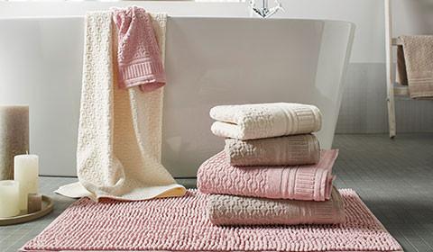 Handtücher in Weiß, Beige und Rosa, gestapelt auf einer rosafarbenen Badematte, von mömax.