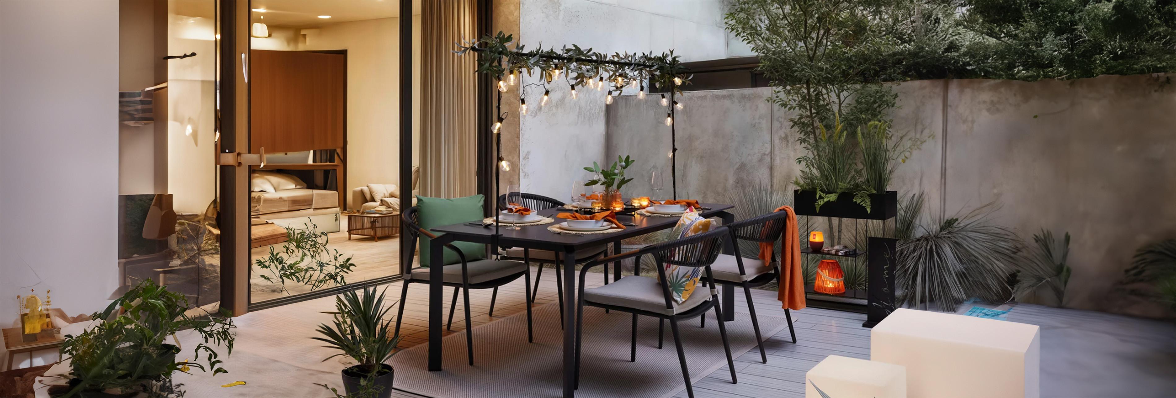 Modern, hangulatos terasz alkonyatkor: kültéri étkezőasztal és székek növényekkel, a jobb előtérben egy természetes kötél/rattan hatású lámpás meleg fénnyel világít, lágy árnyékokat és fényfoltokat vet a fa teraszburkolatra.