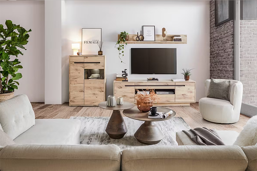 Modern nappali világos fa TV-szekrénnyel és hozzá illő magas szekrénnyel, a falon TV és polc dekorációkkal, elöl fehér kanapé, fotel és dohányzóasztalok.