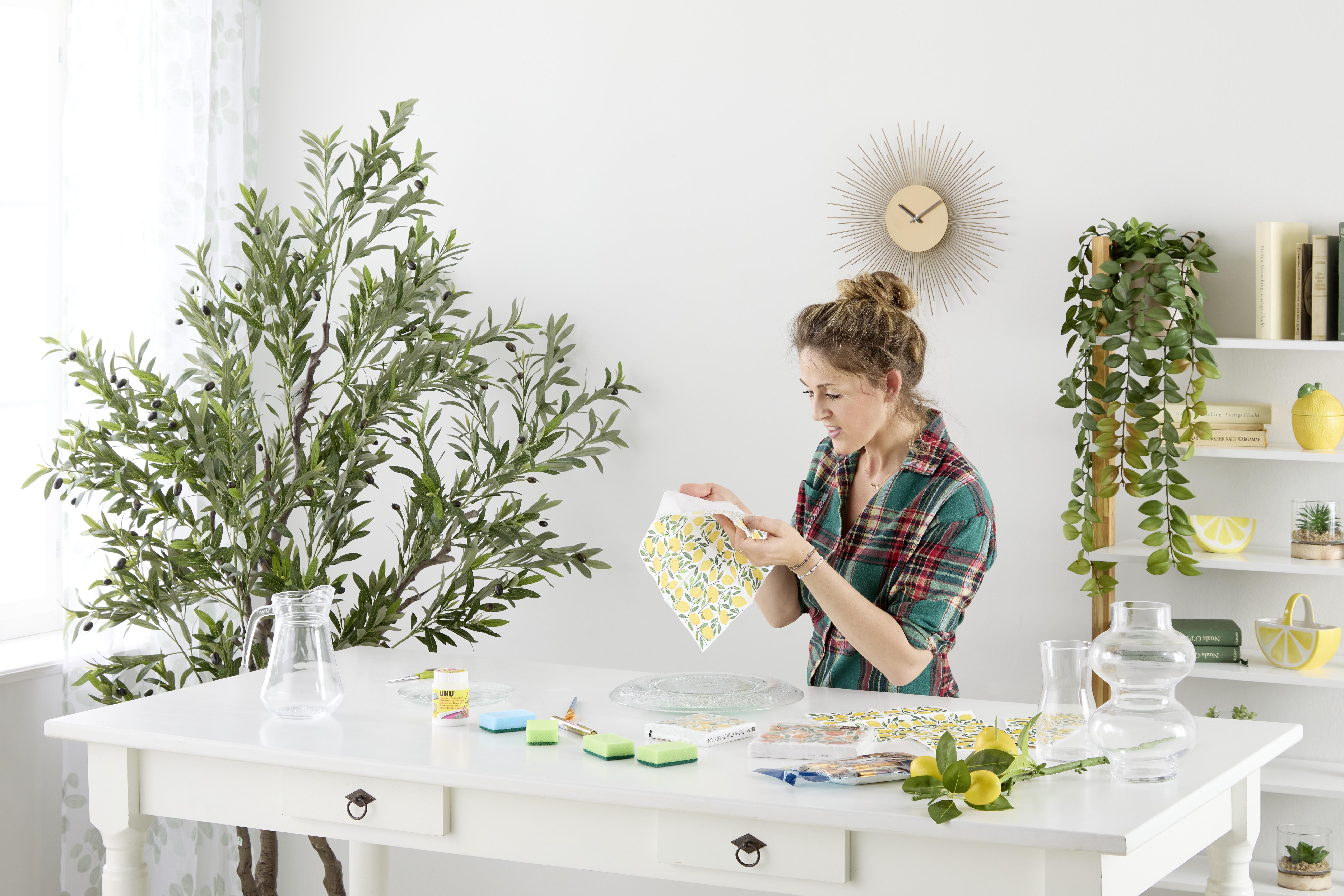 Szalvétatechnikás DIY pillanat: citrommintás szalvétával díszíti az üveg dekorációt világos, növényekkel teli enteriőrben.