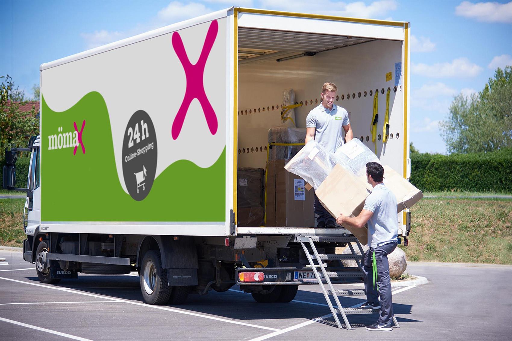 Teherautó nyitott rakterénél két költöztető dolgozó nagy, fóliázott dobozt pakol; a jármű oldalán mömax és 24h online shopping felirat látható.