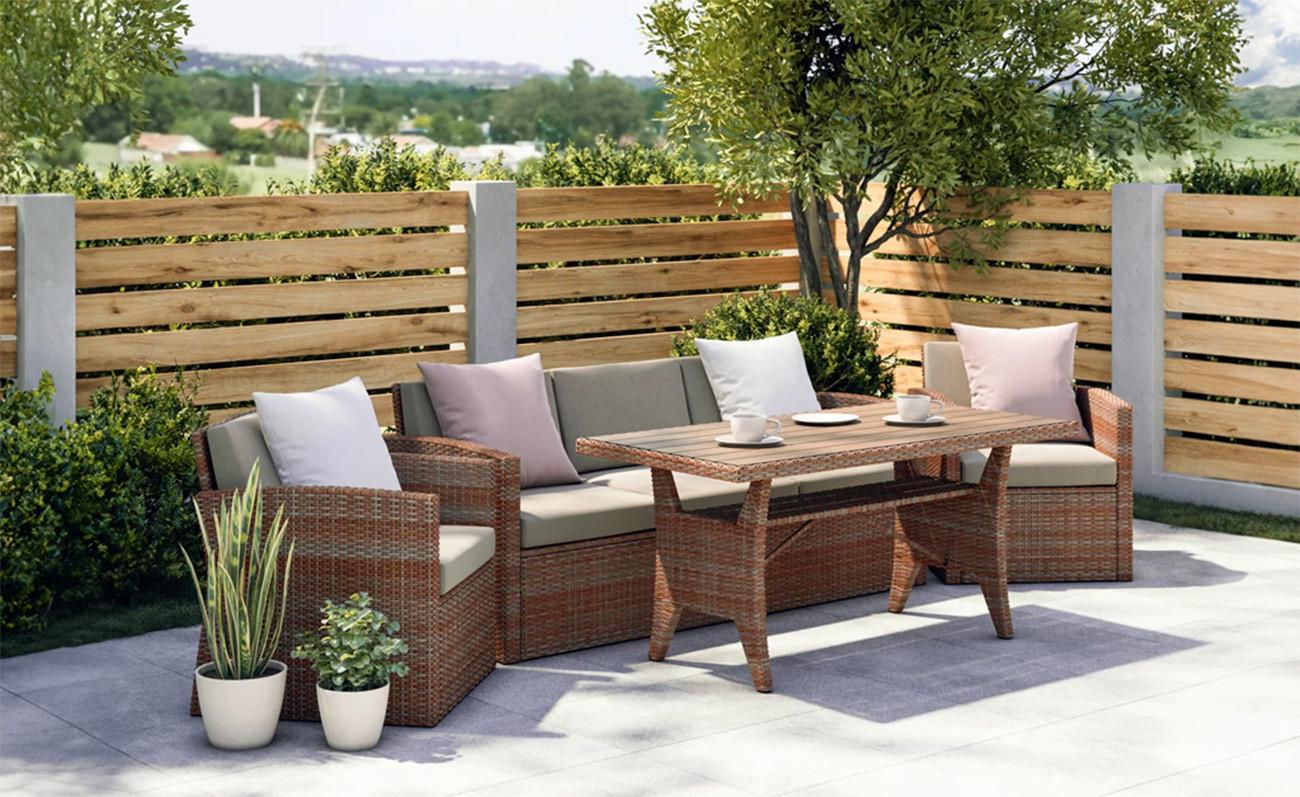 Rattan hatású kerti ülőgarnitúra szürke párnákkal és fehér díszpárnákkal egy modern teraszon. Elöl téglalap alakú asztal csészékkel, háttérben fa kerítés, zöld növények és egy fa.