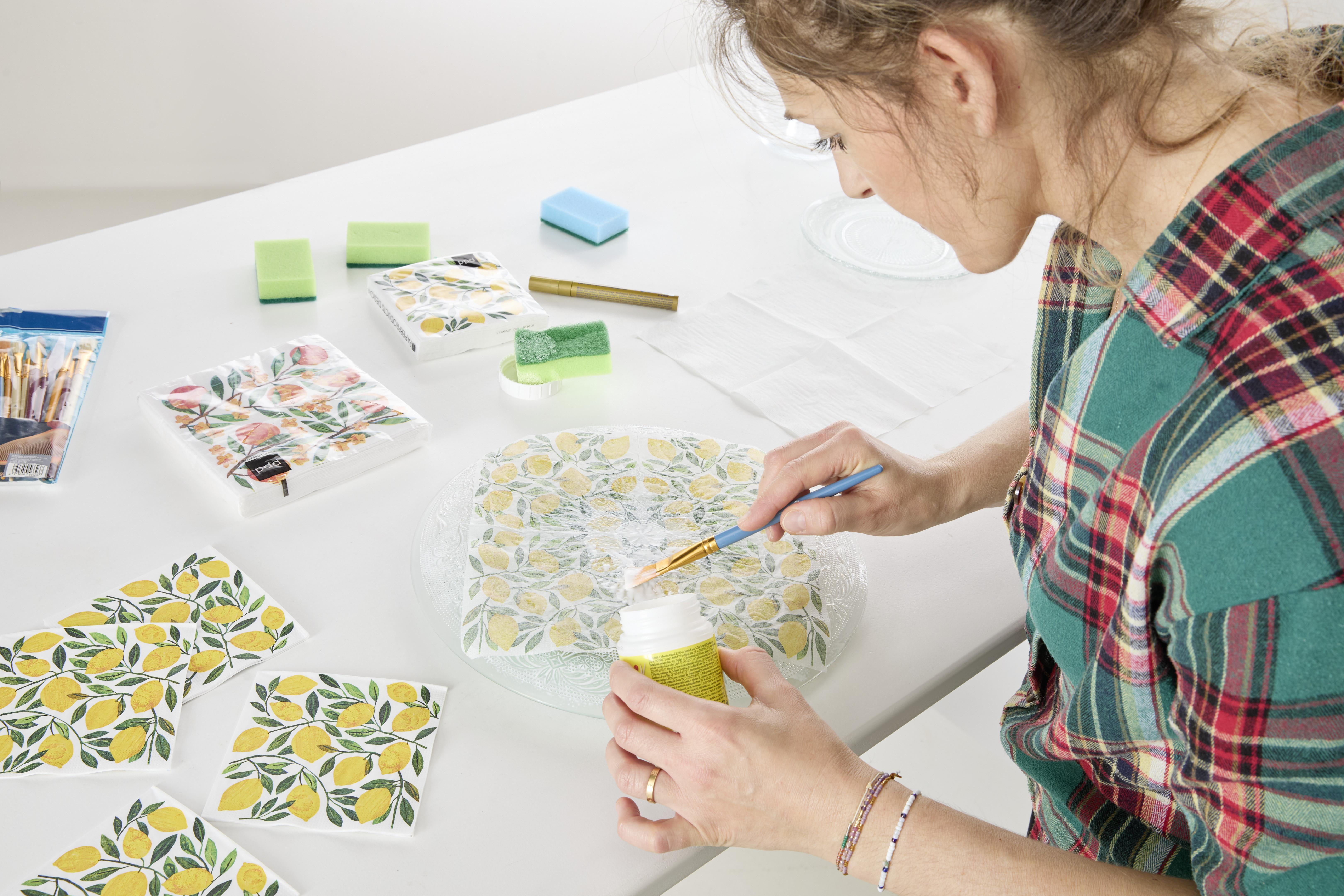 Üvegtányér dekorálása szalvétatechnikával: citrommintás szalvéta rögzítése decoupage ragasztóval és ecsettel, kreatív kellékekkel körülvéve.