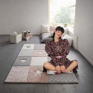 Szürke padlójú szobában patchwork mintás, pasztell gyerekszőnyeg csillag- és szívmotívumokkal; a szőnyegen keresztbe tett lábbal ül egy mosolygó nő, háttérben párnák és játékok.