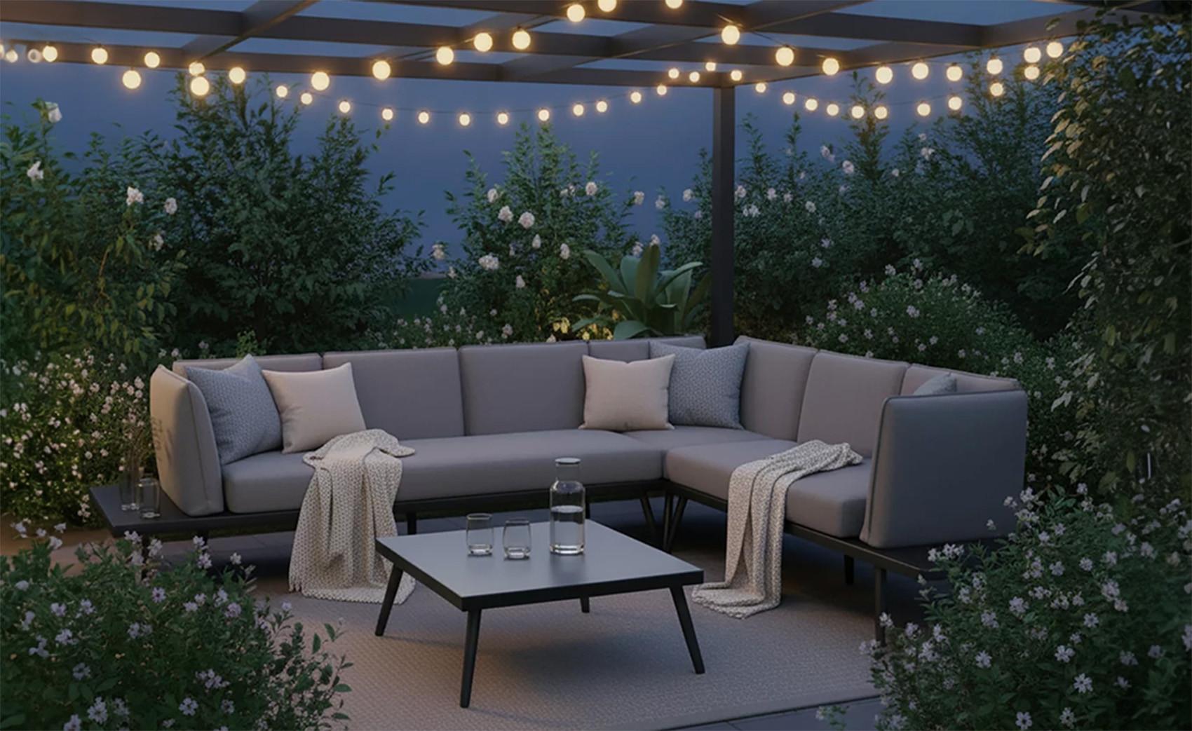 Hangulatos kerti lounge sarokszett szürke párnákkal pergola alatt, fényfüzérekkel megvilágítva, zöld növényekkel körülvéve esti hangulatban.
